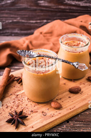 Toffee caramel dessert in jar, sprinkled with grated chocolate. On wooden board, decorated with cinnamon rolls, almonds and star anise. Rustic wooden  Stock Photo