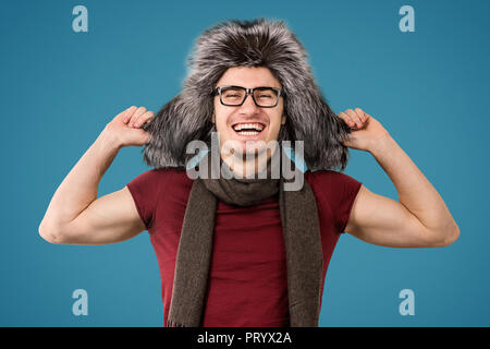 Young man wearing trendy furry winter hat and scarf looking happily at camera on gray background Stock Photo