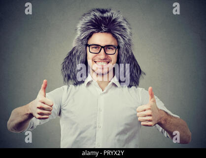 Young man wearing trendy furry winter hat and showing thumb up looking happily at camera on gray background Stock Photo
