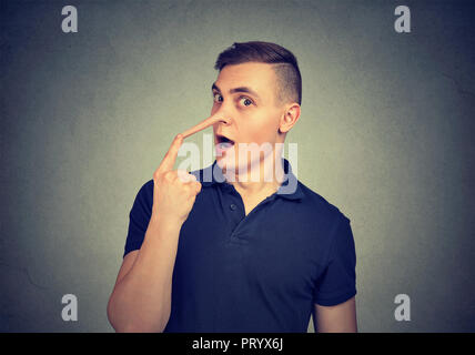 Young man looking surprised at camera touching long fake nose of liar on gray background Stock Photo