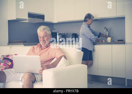 Married adult aged couple at home toghether. women wife wash a mug and dishes and husband man use technology internet laptop to work and read news. da Stock Photo