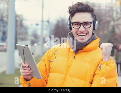 Happy successful man in coat and headphones holding tablet and fist up on street excited with win Stock Photo