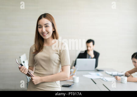 Happy businesswoman standing with arms crossed at office while colleagues discussing in background. Stock Photo
