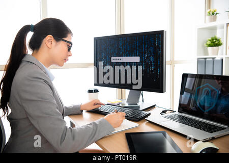 female staff in personal information safety company is sitting at her office desk and writing on notebook while log in Stock Photo