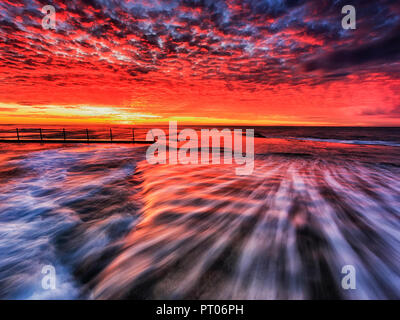Colourful red orange sunrise over sea horizon off Mona Vale beach with rock pool on sandstone plateau in Sydney. Stock Photo