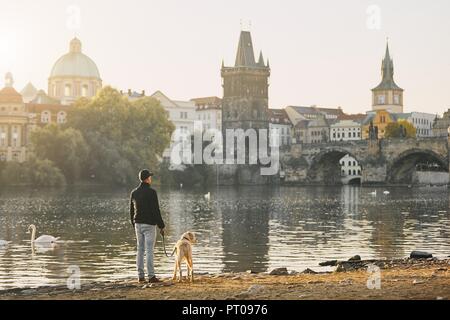 Morning walk with dog. Young man and his labrador retriever on riverbank against Old Town with Charles Bridge in Prague.