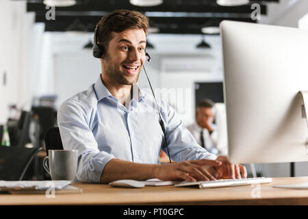 Photo of happy operator man 20s wearing office clothes and headset sitting by computer in call center Stock Photo