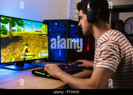 Image of young man playing video games on computer wearing headphones and using backlit colorful keyboard Stock Photo