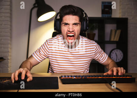 Portrait of angry irritated gamer guy screaming while playing video games on computer wearing headphones and using backlit colorful keyboard Stock Photo