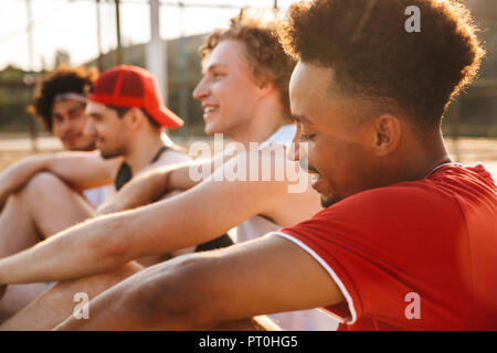 Group of young happy multiethnic men basketball players resting at the sport ground, talking Stock Photo