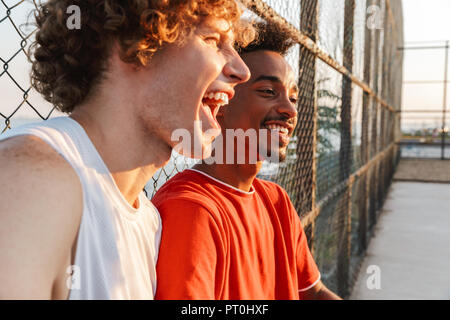 Two young cheerful multiethnic men basketball players resting at the sport ground, talking Stock Photo