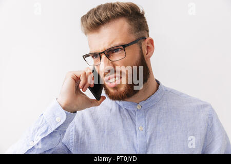 Portrait of an angry young bearded man in eyeglasses standing isolated over white, talking on mobile phone Stock Photo