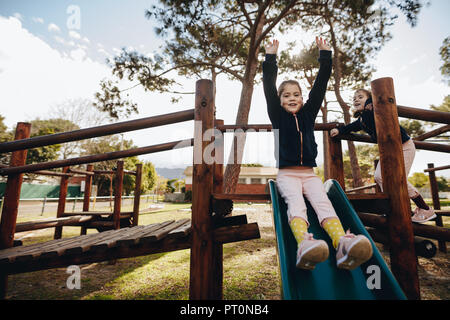 Two active girls playing together outdoors on the slide at the garden. Twin sisters enjoying themselves at the playground. Stock Photo