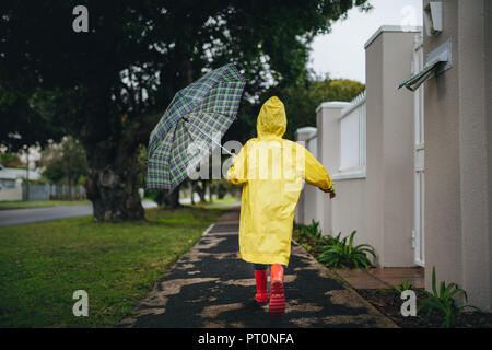 Rear view of little girl running outdoors on a rainy day. Girl wearing raincoat and rubber boots holding umbrella running outside. Stock Photo