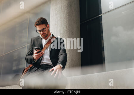 Businessman managing business on the move sitting outdoors. Businessman sitting outside a building looking at his mobile phone. Stock Photo
