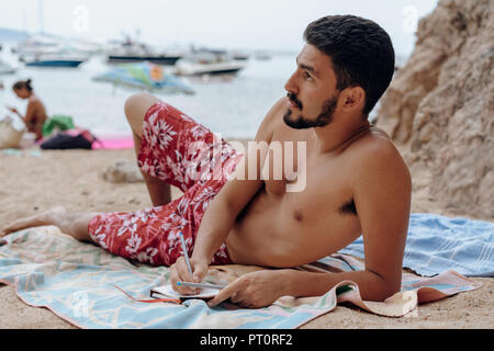 Young man lying on towel at the beach, notebook, thoughtful Stock Photo