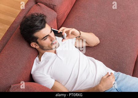 A serious handsome young man in a white tshirt laying on a sofa and talking on his phone. Stock Photo