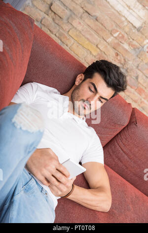 A serious handsome young man in a white tshirt laying on a sofa and using his smartphone. Stock Photo