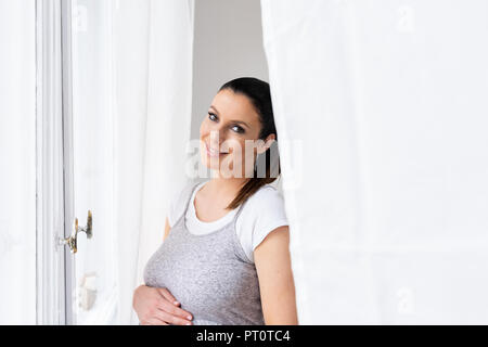 A beautiful young pregnant woman standing next to the window in an elegant grey dress with hands on her belly. Stock Photo