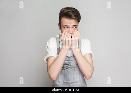 Portrait of nervous young brunette man in casual style with white t-shirt and denim overalls standing and looking away, biting nails with worry face.  Stock Photo