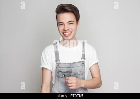 Portrait of happy young brunette man in casual style with white t-shirt and denim overalls standing and looking at camera with toothy smile. indoor st Stock Photo