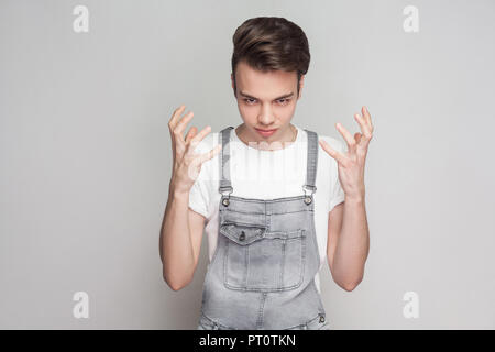 Portrait of angry young brunette man in casual style, white t-shirt and denim overalls standing and looking at camera with raised arms and aggressive.