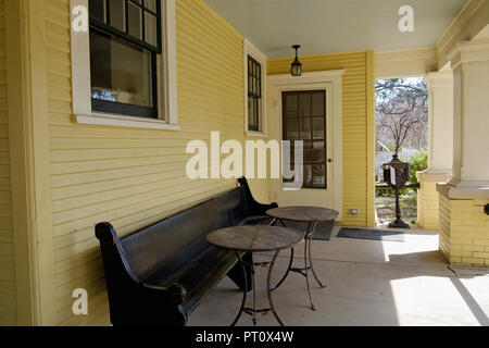 Historic house veransa, long wooden bench & 2 round tables. Yellow shingles & bricks. Dulaney House Chestnut Square Historic Village, McKinney Texas. Stock Photo