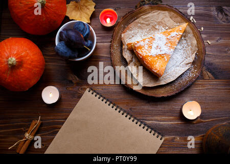 Notebook, pumpkin cheesecake, cooked at home, pumpkin, foliage, table lamp, vanilla on a wooden dark table. Autumn and winter cozy concept. Stock Photo