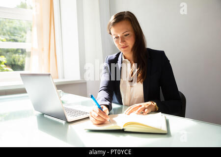 Young Businesswoman Writing Note In Diary With Laptop On Desk Stock Photo