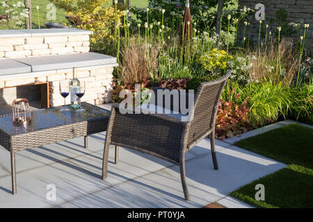 Small urban garden with stone patio with wicker garden patio furniture, coffee table and chair, stone bench, alliums, grasses, spring England UK Stock Photo