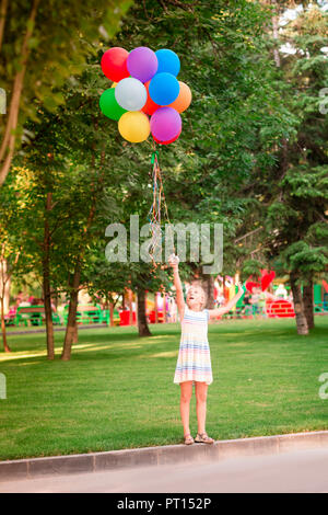 Happy little girl playing with large bunch of helium filled colorful balloons in the park Stock Photo