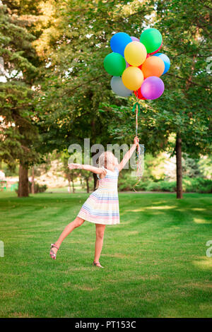 Happy little girl playing with large bunch of helium filled colorful balloons in the park Stock Photo