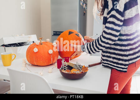 Young woman making jack-o-lantern for halloween on kitchen. Drawing eyes, nose and mouth with pen on pumpkin. Preparing decoration for holiday Stock Photo