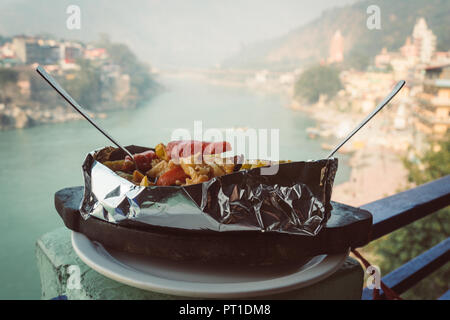 A dish of grilled and stewed vegetables in foil on the background of the river Ganges. Rishikesh India., yoga city India, Gange River Ganga Ram Jhoola Stock Photo