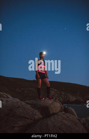 Sportive woman with headlamp standing on rocks in the evening Stock Photo