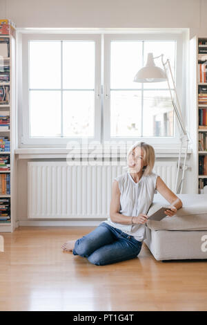 Smiling mature woman sitting on the floor at home holding tablet Stock Photo