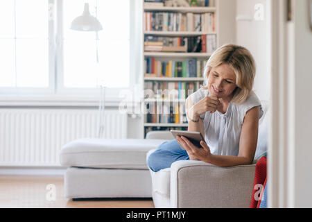 Smiling mature woman lying on couch at home using tablet Stock Photo