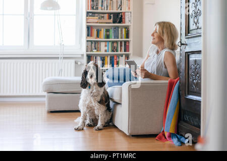 Smiling mature woman with dog lying on couch at home holding tablet Stock Photo