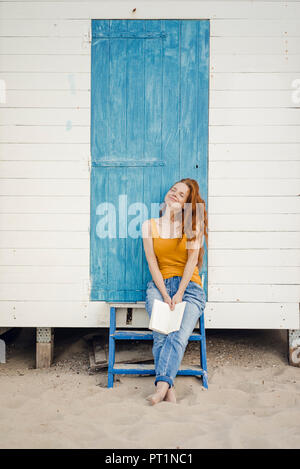 Redheaded woman sitting in front of beach cabin, reading a book Stock Photo