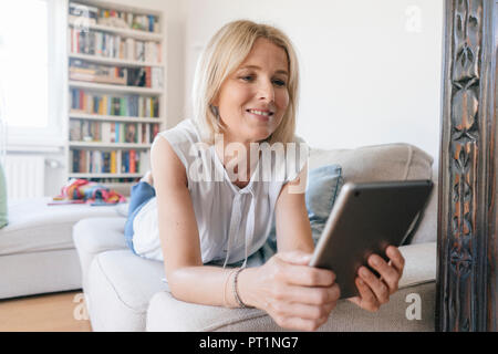 Smiling mature woman lying on couch at home using tablet Stock Photo