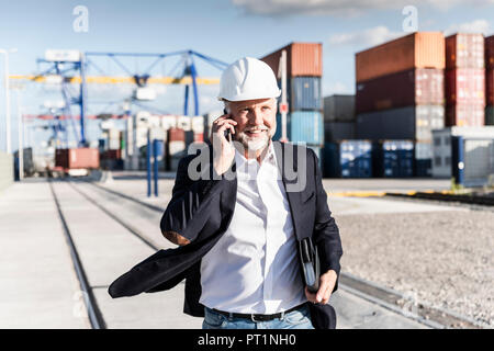 Businessman at cargo harbour, wearing safety helmet, using smartphone Stock Photo