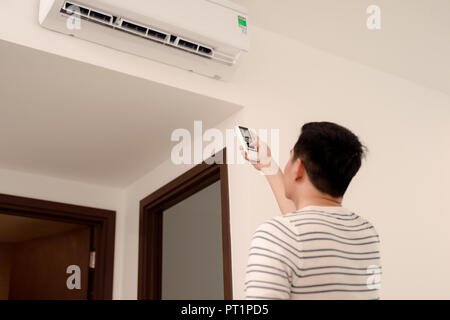 Young man switching on or adjusting the wall mounted air conditioner in the living room with a remote control Stock Photo