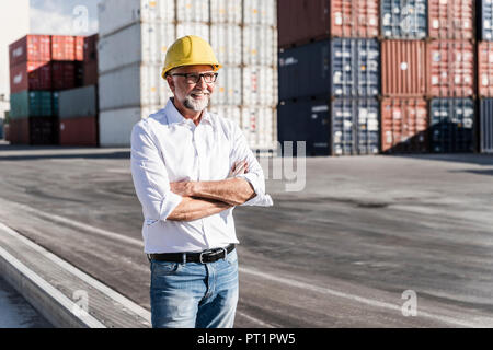 Businessman at cargo harbour, wearing safety helmet, arms crossed Stock Photo
