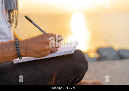 Spain. Man writing on a notebook during sunrise on rocks at the beach Stock Photo
