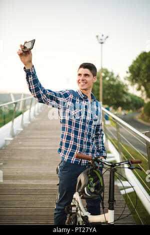 Smiling young man with folding bicycle on a bridge taking a selfie Stock Photo