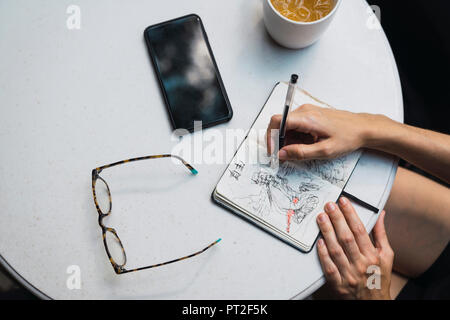 Hands of woman sketching in notebook, glasses, tea and smartphone on table Stock Photo