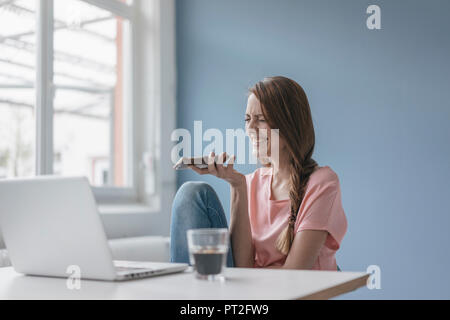 Woman at home sitting at desk with laptop, using smartphone Stock Photo