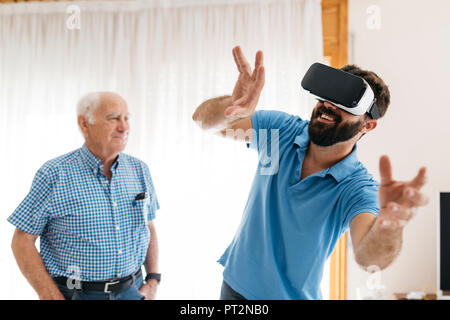 Smiling man using Virtual Reality Glasses at home while his grandfather watching him Stock Photo