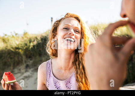 Netherlands, Zandvoort, happy woman eating watermelon looking at man on the beach Stock Photo