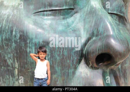 niño posando para la foto con una mano tocándose la cabeza recargado en una escultura verde de una cabeza a gran escala a plana luz del dia Stock Photo